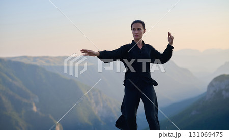 Woman practicing Tai Chi in traditional black outfit on mountain at sunrise, arms outstretched in meditation pose focusing on balance, energy, and mindfulness in nature. 131069475