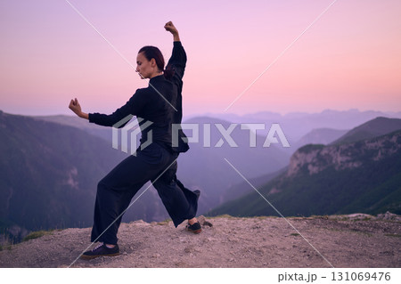 Woman practicing Tai Chi in traditional black outfit on mountain at sunrise, arms outstretched in meditation pose focusing on balance, energy, and mindfulness in nature. 131069476