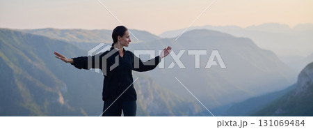 Woman practicing Tai Chi in traditional black outfit on mountain at sunrise, arms outstretched in meditation pose focusing on balance, energy, and mindfulness in nature. 131069484