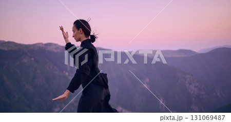 Woman practicing Tai Chi in traditional black outfit on mountain at sunrise, arms outstretched in meditation pose focusing on balance, energy, and mindfulness in nature. 131069487
