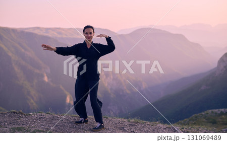 Woman practicing Tai Chi in traditional black outfit on mountain at sunrise, arms outstretched in meditation pose focusing on balance, energy, and mindfulness in nature. 131069489