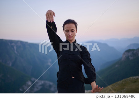 Woman practicing Tai Chi in traditional black outfit on mountain at sunrise, arms outstretched in meditation pose focusing on balance, energy, and mindfulness in nature. 131069490