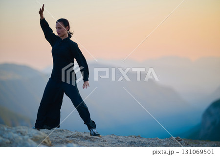 Woman practicing Tai Chi in traditional black outfit on mountain at sunrise, arms outstretched in meditation pose focusing on balance, energy, and mindfulness in nature. 131069501
