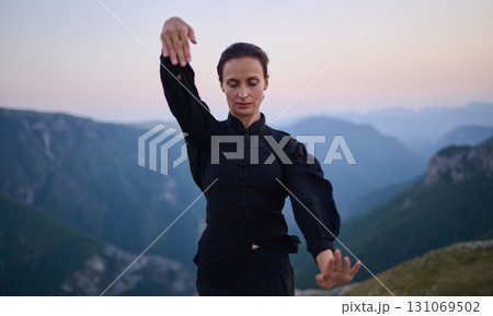 Woman practicing Tai Chi in traditional black outfit on mountain at sunrise, arms outstretched in meditation pose focusing on balance, energy, and mindfulness in nature. Woman practicing Tai Chi in traditional black outfit on mountain at sunrise, arms outstretched in meditation pose focusing on balance, energy, and mindfulness in nature. 131069502