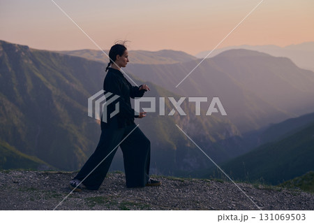 Woman practicing Tai Chi in traditional black outfit on mountain at sunrise, arms outstretched in meditation pose focusing on balance, energy, and mindfulness in nature. Woman practicing Tai Chi in traditional black outfit on mountain at sunrise, arms outstretched in meditation pose focusing on balance, energy, and mindfulness in nature. 131069503