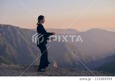 Woman practicing Tai Chi in traditional black outfit on mountain at sunrise, arms outstretched in meditation pose focusing on balance, energy, and mindfulness in nature. 131069504
