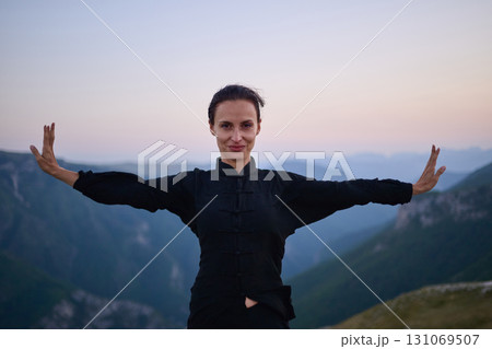 Woman practicing Tai Chi in traditional black outfit on mountain at sunrise, arms outstretched in meditation pose focusing on balance, energy, and mindfulness in nature. 131069507