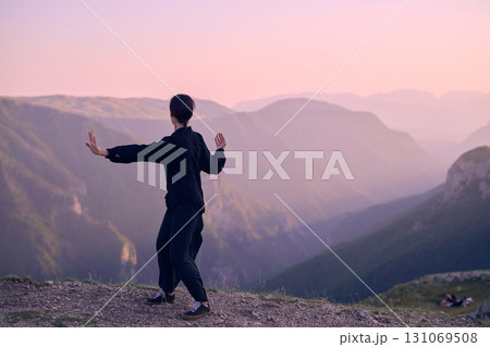 Woman practicing Tai Chi in traditional black outfit on mountain at sunrise, arms outstretched in meditation pose focusing on balance, energy, and mindfulness in nature. 131069508