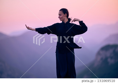 Woman practicing Tai Chi in traditional black outfit on mountain at sunrise, arms outstretched in meditation pose focusing on balance, energy, and mindfulness in nature. 131069510