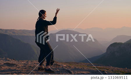 Woman practicing Tai Chi in traditional black outfit on mountain at sunrise, arms outstretched in meditation pose focusing on balance, energy, and mindfulness in nature. 131069511