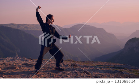 Woman practicing Tai Chi in traditional black outfit on mountain at sunrise, arms outstretched in meditation pose focusing on balance, energy, and mindfulness in nature. 131069514