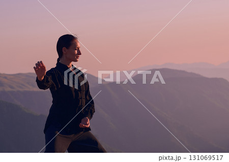 Woman practicing Tai Chi in traditional black outfit on mountain at sunrise, arms outstretched in meditation pose focusing on balance, energy, and mindfulness in nature. 131069517