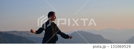 Woman practicing Tai Chi in traditional black outfit on mountain at sunrise, arms outstretched in meditation pose focusing on balance, energy, and mindfulness in nature. 131069529