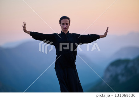 Woman practicing Tai Chi in traditional black outfit on mountain at sunrise, arms outstretched in meditation pose focusing on balance, energy, and mindfulness in nature. 131069530
