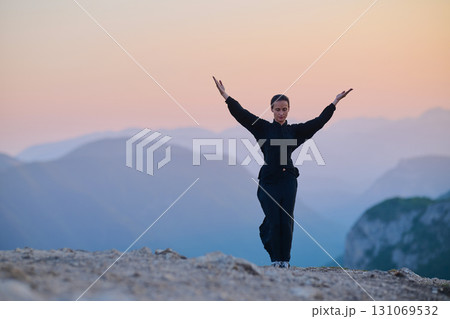 Woman practicing Tai Chi in traditional black outfit on mountain at sunrise, arms outstretched in meditation pose focusing on balance, energy, and mindfulness in nature. 131069532