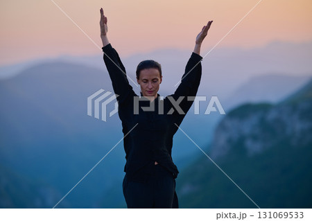 Woman practicing Tai Chi in traditional black outfit on mountain at sunrise, arms outstretched in meditation pose focusing on balance, energy, and mindfulness in nature. 131069533