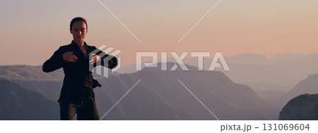 Woman practicing Tai Chi in traditional black outfit on mountain at sunrise, arms outstretched in meditation pose focusing on balance, energy, and mindfulness in nature. 131069604