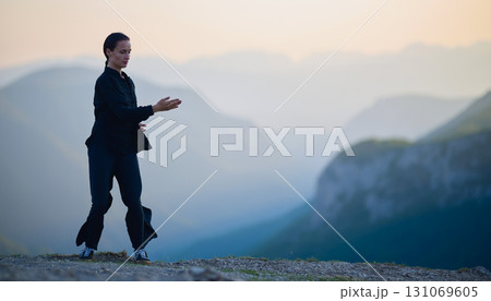 Woman practicing Tai Chi in traditional black outfit on mountain at sunrise, arms outstretched in meditation pose focusing on balance, energy, and mindfulness in nature. 131069605