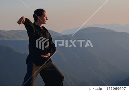 Woman practicing Tai Chi in traditional black outfit on mountain at sunrise, arms outstretched in meditation pose focusing on balance, energy, and mindfulness in nature. 131069607