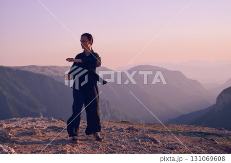 Woman practicing Tai Chi in traditional black outfit on mountain at sunrise, arms outstretched in meditation pose focusing on balance, energy, and mindfulness in nature. 131069608