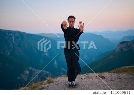 Woman practicing Tai Chi in traditional black outfit on mountain at sunrise, arms outstretched in meditation pose focusing on balance, energy, and mindfulness in nature. Woman practicing Tai Chi in traditional black outfit on mountain at sunrise, arms outstretched in meditation pose focusing on balance, energy, and mindfulness in nature. 131069611