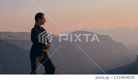 Woman practicing Tai Chi in traditional black outfit on mountain at sunrise, arms outstretched in meditation pose focusing on balance, energy, and mindfulness in nature. 131069630