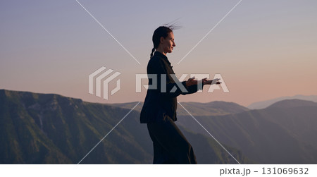 Woman practicing Tai Chi in traditional black outfit on mountain at sunrise, arms outstretched in meditation pose focusing on balance, energy, and mindfulness in nature. 131069632