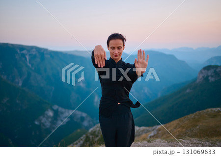 Woman practicing Tai Chi in traditional black outfit on mountain at sunrise, arms outstretched in meditation pose focusing on balance, energy, and mindfulness in nature. 131069633