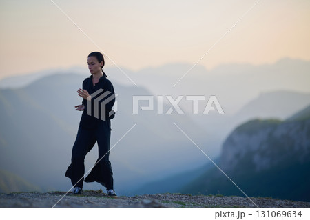 Woman practicing Tai Chi in traditional black outfit on mountain at sunrise, arms outstretched in meditation pose focusing on balance, energy, and mindfulness in nature. Woman practicing Tai Chi in traditional black outfit on mountain at sunrise, arms outstretched in meditation pose focusing on balance, energy, and mindfulness in nature. 131069634