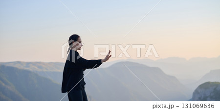 Woman practicing Tai Chi in traditional black outfit on mountain at sunrise, arms outstretched in meditation pose focusing on balance, energy, and mindfulness in nature. 131069636