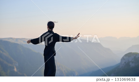 Woman practicing Tai Chi in traditional black outfit on mountain at sunrise, arms outstretched in meditation pose focusing on balance, energy, and mindfulness in nature. 131069638