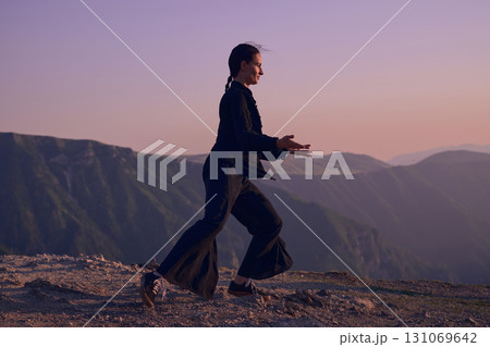 Woman practicing Tai Chi in traditional black outfit on mountain at sunrise, arms outstretched in meditation pose focusing on balance, energy, and mindfulness in nature. Woman practicing Tai Chi in traditional black outfit on mountain at sunrise, arms outstretched in meditation pose focusing on balance, energy, and mindfulness in nature. 131069642