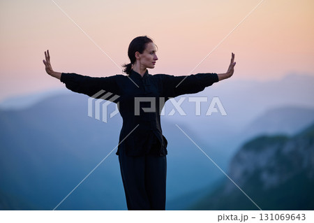 Woman practicing Tai Chi in traditional black outfit on mountain at sunrise, arms outstretched in meditation pose focusing on balance, energy, and mindfulness in nature. 131069643