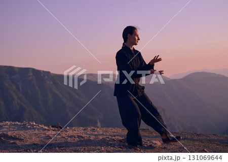 Woman practicing Tai Chi in traditional black outfit on mountain at sunrise, arms outstretched in meditation pose focusing on balance, energy, and mindfulness in nature. 131069644