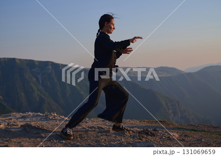 Woman practicing Tai Chi in traditional black outfit on mountain at sunrise, arms outstretched in meditation pose focusing on balance, energy, and mindfulness in nature. 131069659