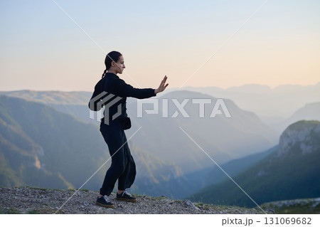 Woman practicing Tai Chi in traditional black outfit on mountain at sunrise, arms outstretched in meditation pose focusing on balance, energy, and mindfulness in nature. 131069682