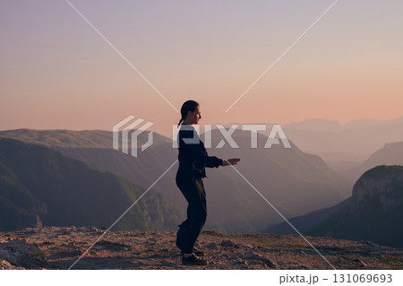 Woman practicing Tai Chi in traditional black outfit on mountain at sunrise, arms outstretched in meditation pose focusing on balance, energy, and mindfulness in nature. 131069693