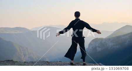 Woman practicing Tai Chi in traditional black outfit on mountain at sunrise, arms outstretched in meditation pose focusing on balance, energy, and mindfulness in nature. 131069708