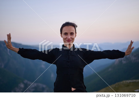 Woman practicing Tai Chi in traditional black outfit on mountain at sunrise, arms outstretched in meditation pose focusing on balance, energy, and mindfulness in nature. Woman practicing Tai Chi in traditional black outfit on mountain at sunrise, arms outstretched in meditation pose focusing on balance, energy, and mindfulness in nature. 131069710