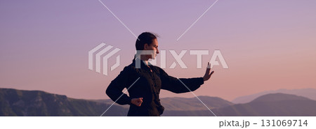 Woman practicing Tai Chi in traditional black outfit on mountain at sunrise, arms outstretched in meditation pose focusing on balance, energy, and mindfulness in nature. 131069714