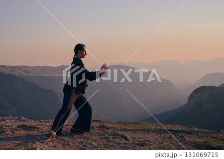 Woman practicing Tai Chi in traditional black outfit on mountain at sunrise, arms outstretched in meditation pose focusing on balance, energy, and mindfulness in nature. 131069715