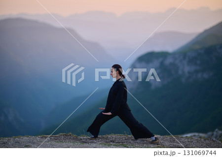 Woman practicing Tai Chi in traditional black outfit on mountain at sunrise, arms outstretched in meditation pose focusing on balance, energy, and mindfulness in nature. 131069744