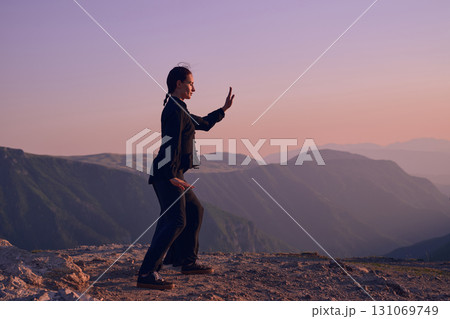 Woman practicing Tai Chi in traditional black outfit on mountain at sunrise, arms outstretched in meditation pose focusing on balance, energy, and mindfulness in nature. Woman practicing Tai Chi in traditional black outfit on mountain at sunrise, arms outstretched in meditation pose focusing on balance, energy, and mindfulness in nature. 131069749