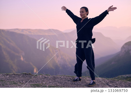 Woman practicing Tai Chi in traditional black outfit on mountain at sunrise, arms outstretched in meditation pose focusing on balance, energy, and mindfulness in nature. 131069750