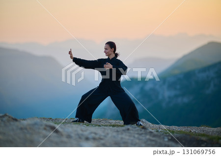 Woman practicing Tai Chi in traditional black outfit on mountain at sunrise, arms outstretched in meditation pose focusing on balance, energy, and mindfulness in nature. 131069756