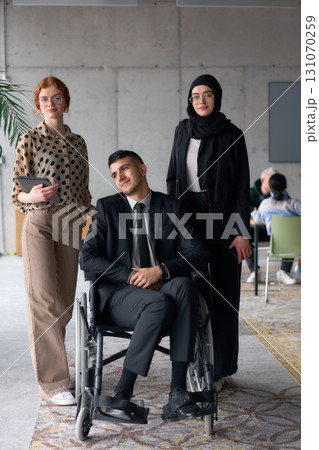 A diverse business team poses confidently, including a smiling director seated in a wheelchair, flanked by two professional female colleagues, one wearing a hijab and the other holding a tablet, in a 131070259