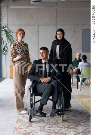 A diverse business team poses confidently, including a smiling director seated in a wheelchair, flanked by two professional female colleagues, one wearing a hijab and the other holding a tablet, in a 131070260