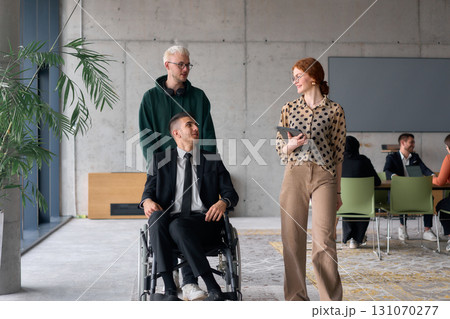 A diverse team of business colleagues, including a director in a wheelchair, engages in a collaborative discussion as they move together through a modern office space A diverse team of business colleagues, including a director in a wheelchair, engages in a collaborative discussion as they move together through a modern office space 131070277