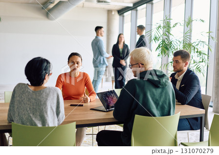 A diverse team of business professionals engaged in a discussion around a conference table in an office, while their colleagues collaborate in the background A diverse team of business professionals engaged in a discussion around a conference table in an office, while their colleagues collaborate in the background 131070279