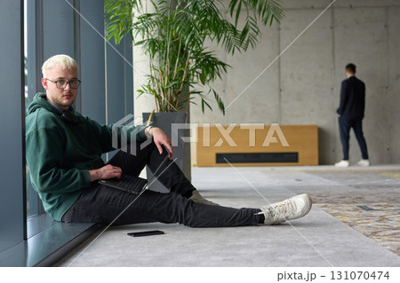 A casual young professional sitting by a large window in a modern office, smiling while working on a laptop, with colleagues interacting in the blurred background. 131070474